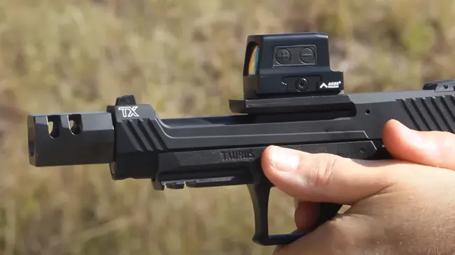 Close-up of a person's hand holding a Taurus TX22 Competition SCR pistol with a mounted red dot sight, against a blurred natural background.