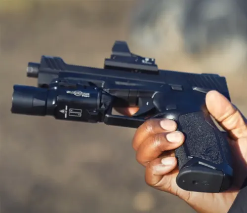A close-up of a hand gripping a Taurus TX22 Competition SCR pistol with attached suppressor and red dot sight, against a blurred natural background.