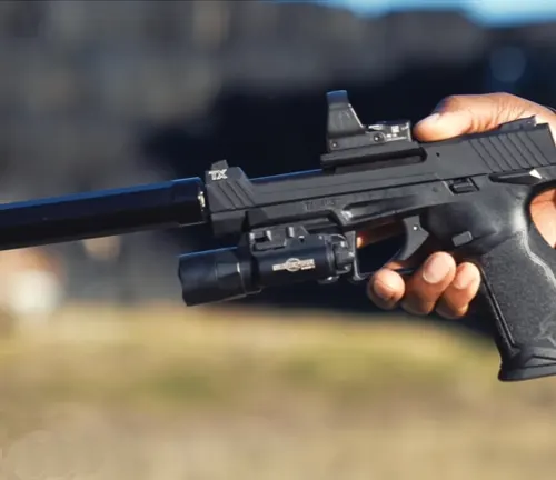 A Taurus TX22 Competition SCR pistol with a suppressor and reflex sight, held in a shooter's hand against a blurred outdoor background.