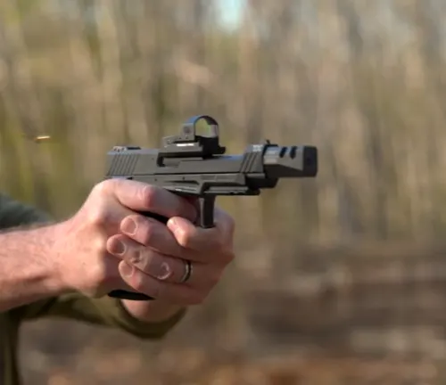 A person's hands steadying a Taurus TX22 Competition SCR pistol with an attached red dot sight, with a spent cartridge case being ejected, against a blurred woodland background.