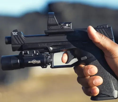 A close-up of a Taurus TX22 Competition SCR pistol with a mounted red dot sight being aimed, held securely in a shooter's hand against a blurred natural background.