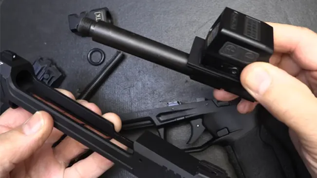 Hands disassembling a Taurus TX22 Competition SCR, showing the detached slide with a mounted red dot sight, barrel, and recoil spring on a textured surface.