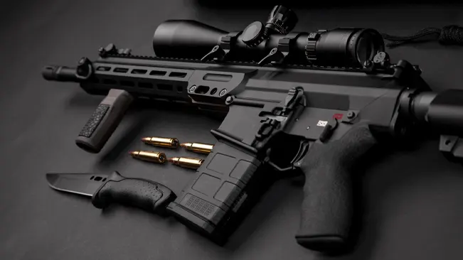 A black tactical rifle with a scope, additional rail accessories, and a large magazine is displayed on a dark surface. Next to it, there are loose bullets, a knife with a black handle, and an additional magazine. The setup suggests a focus on the equipment's details and readiness for use.
