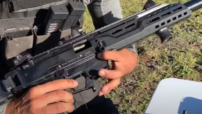 A person is holding a black tactical rifle with both hands, fingers near the trigger guard, with a red dot sight mounted on top. The rifle is partly disassembled or opened for maintenance or reloading, with a grassy outdoor area in the background.