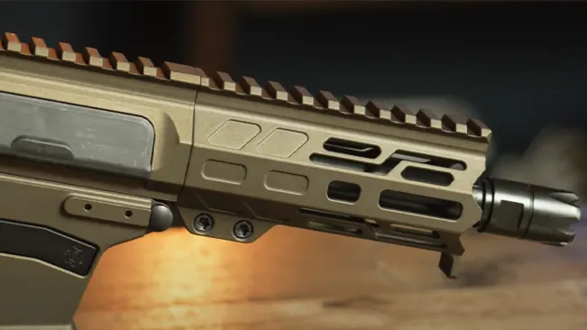 Close-up of the front part of a tan tactical rifle showing the barrel, muzzle device, and part of the handguard with rail attachments on a blurred background.