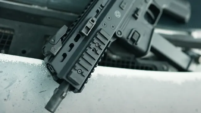 A black tactical rifle with picatinny rails and a suppressor is placed diagonally across a snowy surface, with a metallic structure in the background.