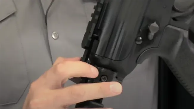 A person's hand adjusting the bolt attached to the undercarriage of a black tactical rifle, against a grey shirt background.