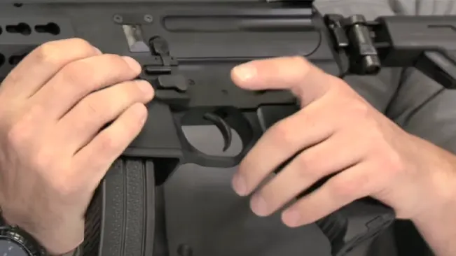 Two hands working on the upper receiver of a disassembled black tactical rifle, with a focus on the trigger and magazine area.