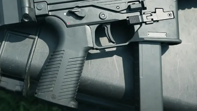 Close-up view of a black firearm's grip, trigger, and lower receiver with an attached magazine and a visible safety selector switch with a red indicator.
