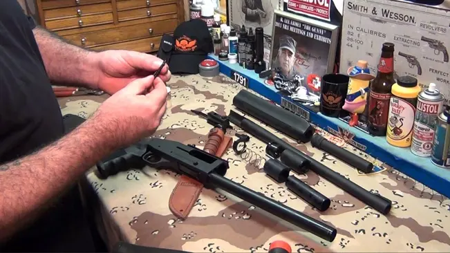 Disassembled Mossberg 930 shotgun parts laid out on a camouflaged mat with a person cleaning a small component at a workbench with gun maintenance tools and products