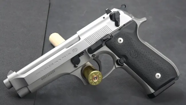 Stainless steel Beretta 92FS INOX with black grips and a chambered round, resting on a dark surface.