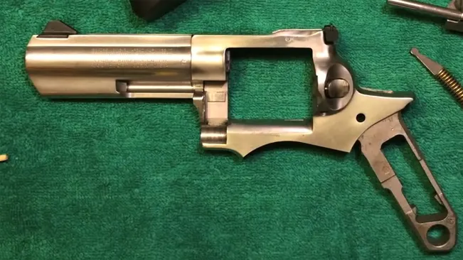 Disassembled stainless steel frame of a Ruger GP100 revolver on a green mat with parts scattered around.
