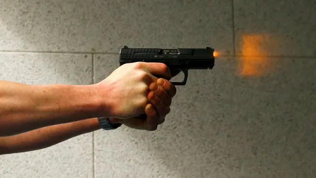 Hand firing a Walther P99 pistol with visible muzzle flash against a concrete wall background