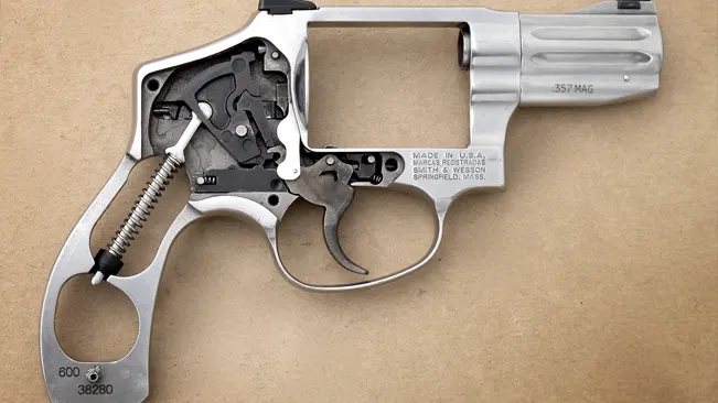 Disassembled Smith & Wesson stainless steel revolver frame with exposed trigger mechanism and .357 MAG inscription, on a beige background.