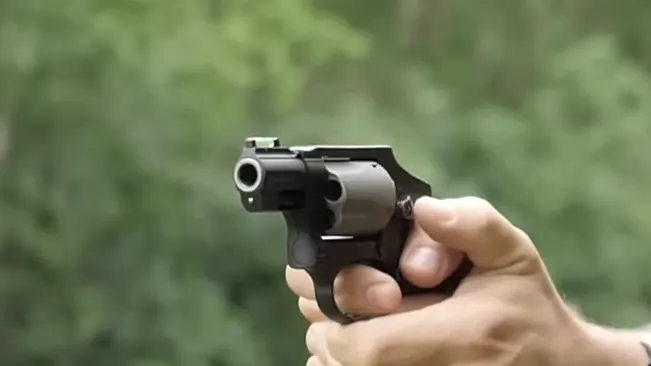 Hand holding a Smith & Wesson J-Frame 340 PD revolver aimed forward, with a blurred green background.