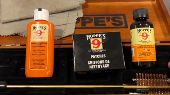 Gun cleaning supplies including Hoppe's 9 lubricating oil, solvent, patches, and cleaning rods on a wooden table.