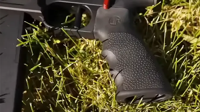Close-up of a textured pistol grip with a red safety indicator, lying on grass.