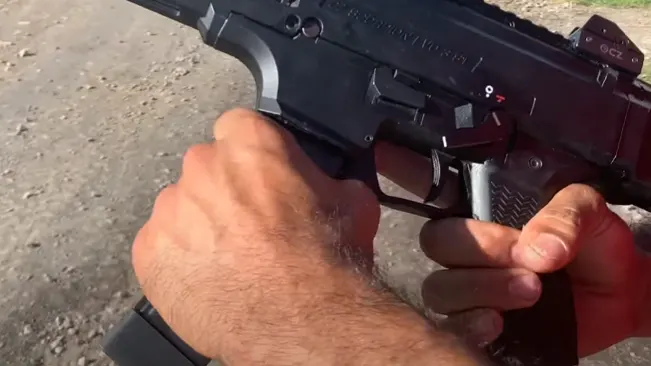 A person's hand is gripping the handle and trigger of a black pistol with a visible red dot sight and a portion of a magazine. The focus is on the action of the hand on the firearm, with a gravel background suggesting an outdoor setting.