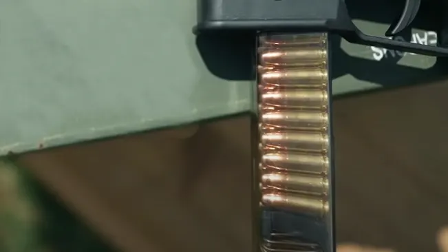 Close-up of a transparent firearm magazine loaded with copper-colored bullets, attached to the black handle of a gun, with a blurred green background.