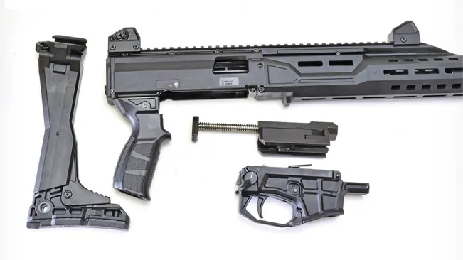 A disassembled black tactical rifle is laid out on a white background. The components include the main body with the barrel, a bipod, a pistol grip, a magazine, a spring, and a lower receiver with a trigger assembly. The image is arranged to show all parts clearly for identification or assembly purposes.