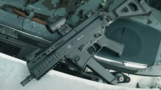 A black tactical rifle with a red dot sight, resting on a textured metallic surface, possibly part of a vehicle or machinery, with hues of blue and rust.