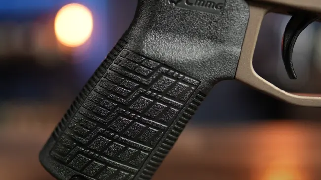 Close-up of the textured grip of a black pistol with branding, against a blurred background with warm light.