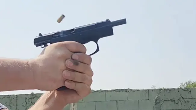 A photo capturing a handgun mid-firing, with a cartridge casing ejected into the air and the shooter's hands gripping the firearm firmly. The background shows a clear sky and the top of a concrete wall.