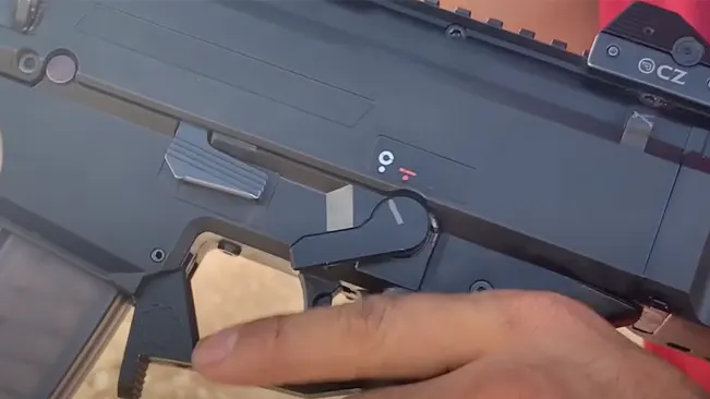 A person's hand is inserting a beige magazine into the magazine well of a black rifle with a visible serial number and a red dot sight on top. The focus is on the action of reloading the weapon.