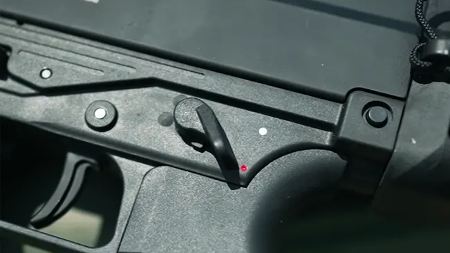 Close-up of a black firearm's safety selector switch with a red dot indicating the fire mode, against a blurred background.