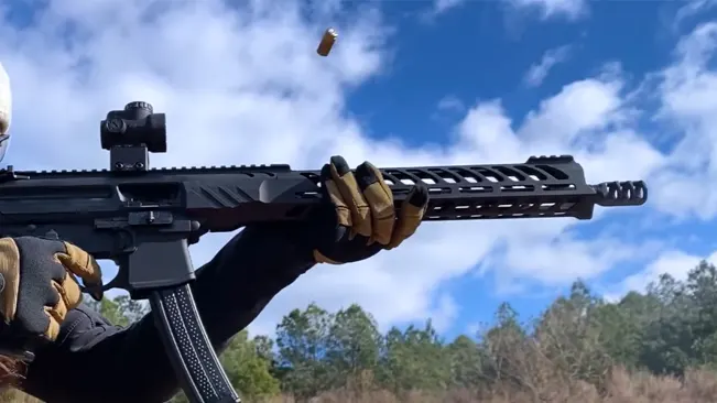 A person wearing gloves is holding a black tactical rifle outdoors, with a brass cartridge casing being ejected into the blue sky with clouds.