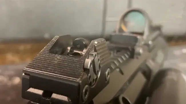This is a close-up image of the rear iron sight on a rifle, with a blurred red dot sight in the background, emphasizing the alignment and focus on precision in shooting.