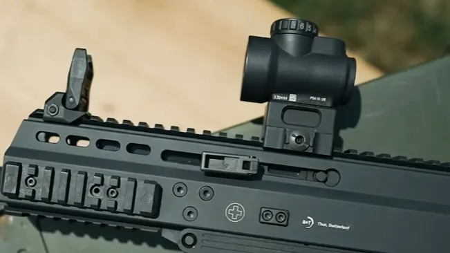 A close-up of a black rifle's upper receiver with a mounted red dot optic and flip-up iron sight, set against a natural outdoor background.