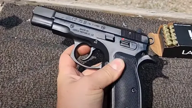 A photo of a person's hand holding a CZ 75 B 9mm handgun with a box of ammunition visible in the background, placed on a textured surface.