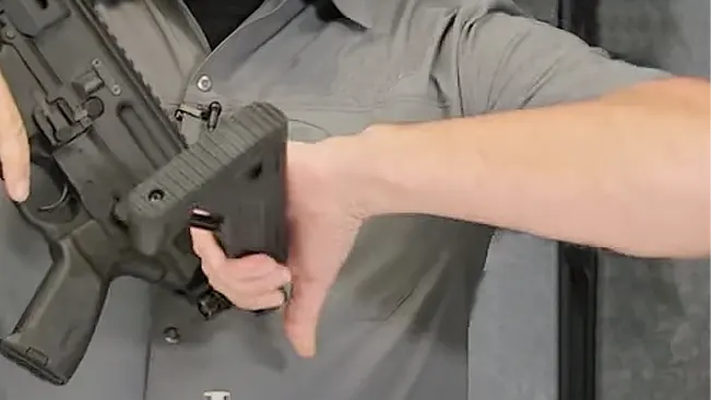 A person is inserting a magazine into the magwell of a black AR-15 style rifle against a grey background.