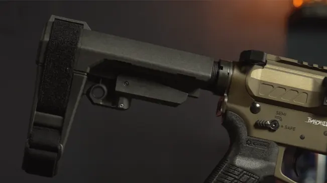 Close-up of a tan and black firearm's rear, featuring an adjustable stock and textured pistol grip, with the 'BANSHEE' logo and fire selector marked 'SEMI' and 'SAFE', against a blurred background.