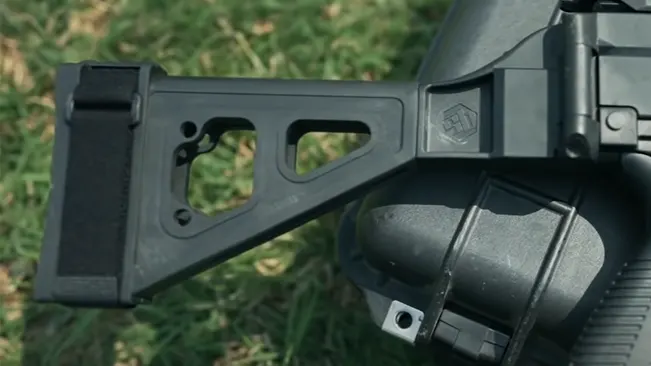 A close-up of the stock and ear cup of hearing protection on a black rifle against a natural green grass background.