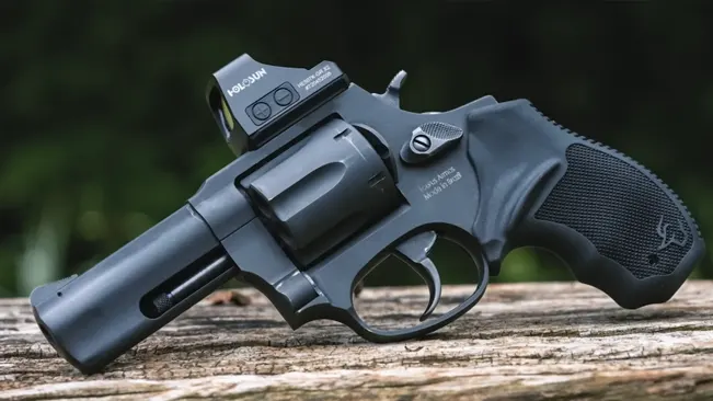 Alt text: "Taurus 856 TORO revolver with a matte black finish and a red dot sight, resting on a wooden log.