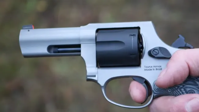 Hand holding a Taurus Defender 856 revolver with a matte stainless finish and red front sight, outdoors.